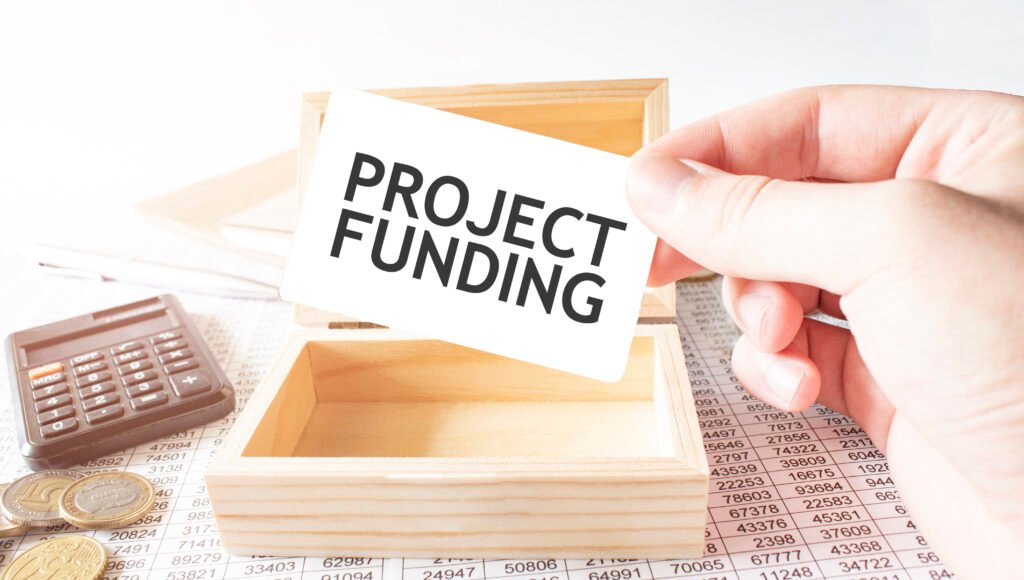 Hand holding a "PROJECT FUNDING" card, with a calculator, coins, and financial documents in the background.
