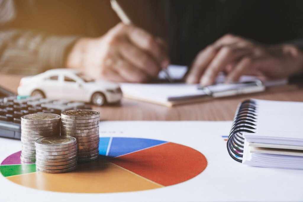 Stacks of coins placed on a colorful pie chart, with a calculator, a miniature car, and a person writing in a notebook in the background. The scene represents financial management, budgeting, or insurance planning.