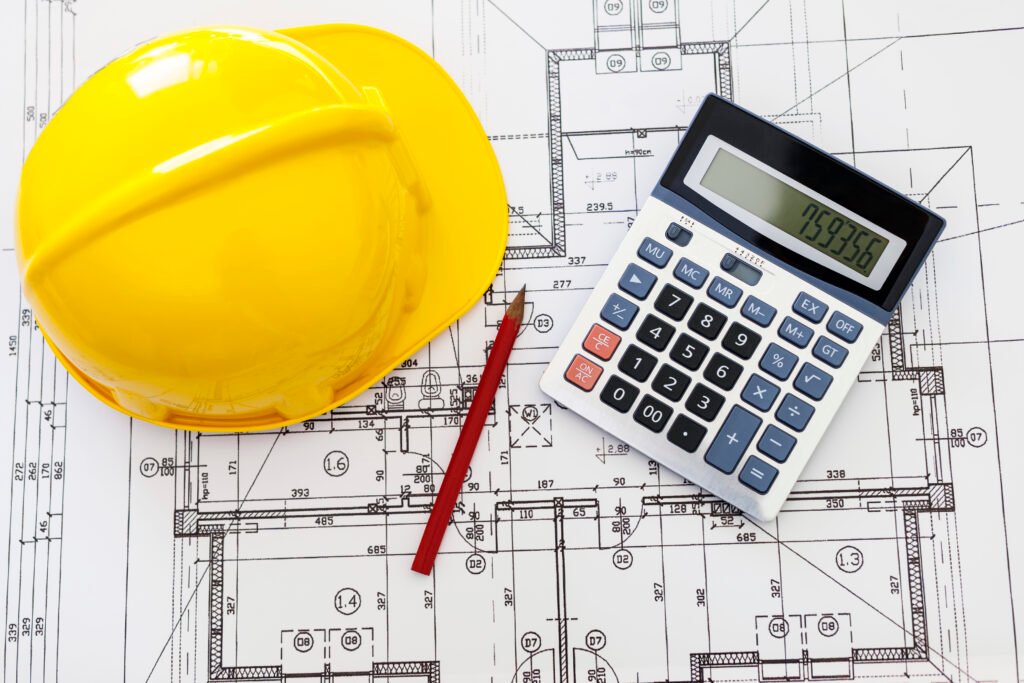 A yellow hard hat, red pencil, and calculator placed on architectural blueprints.
The image symbolizes cost estimation, budgeting, and planning in construction projects.