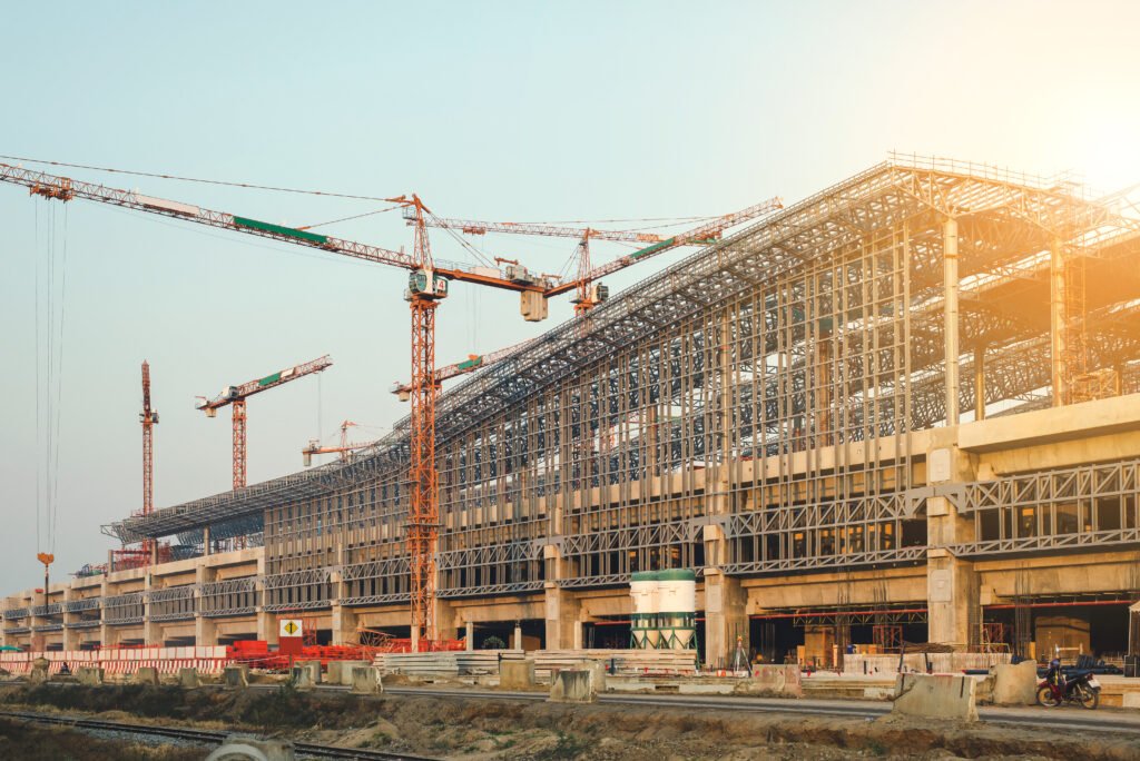 A large construction site with multiple cranes working on a massive steel-framed structure. The site shows ongoing work with building materials and equipment scattered around.