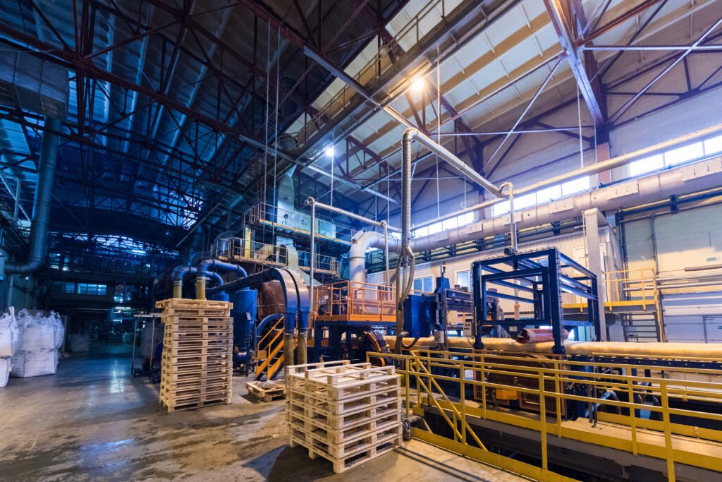 nterior view of a large industrial factory with machinery, pipes, and equipment used in the production process. Stacks of wooden pallets and large bags of raw materials are visible.