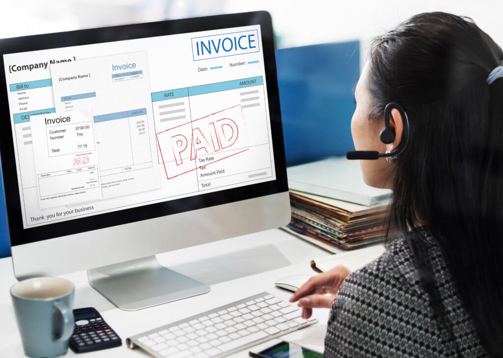 A woman wearing a headset works on a computer screen displaying invoices with a large "PAID" stamp. The workspace includes a keyboard, calculator, coffee cup, and neatly stacked documents, illustrating digital financial transactions.