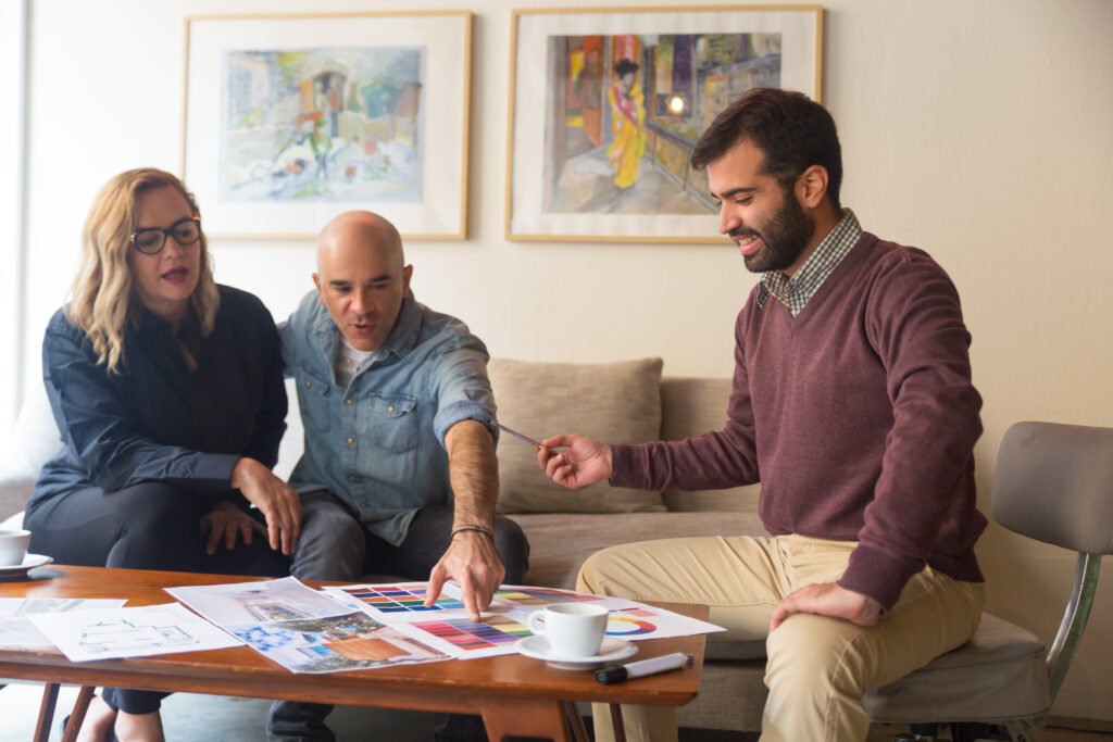 A middle-aged couple and a man sit around a table discussing a renovation project. They are looking at color swatches, floor plans, and design ideas with coffee cups on the table.