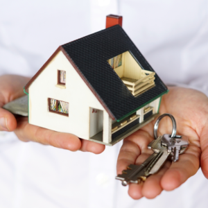 A person holding a small model house in one hand and a set of keys in the other, representing homeownership or real estate transaction.