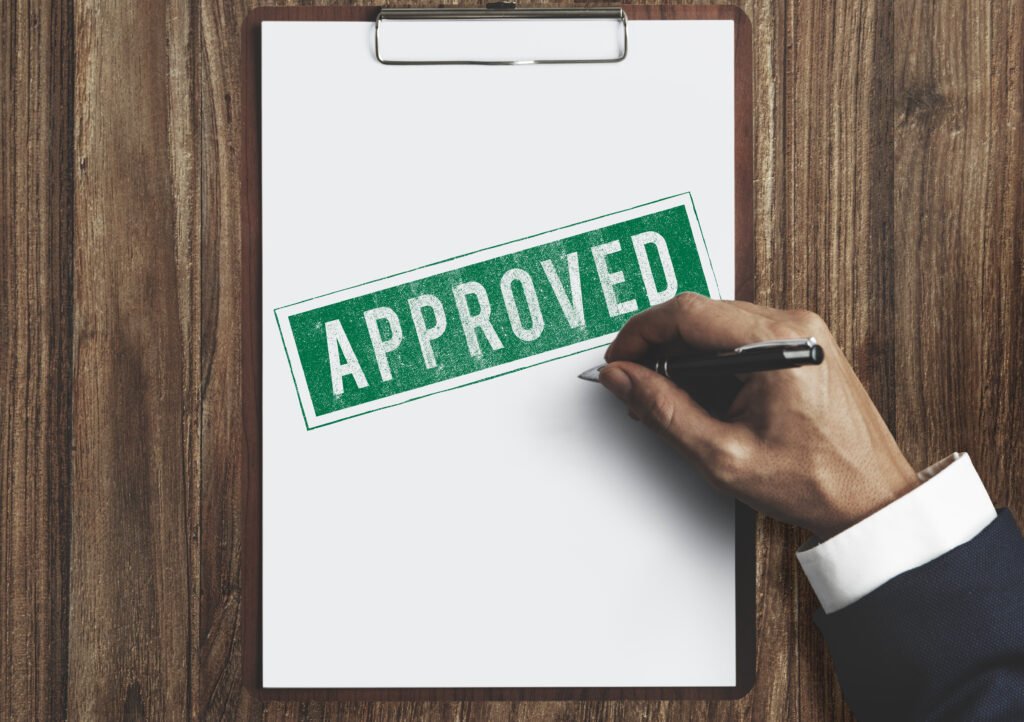 A hand in a dark suit is holding a pen next to a clipboard with a white sheet of paper that has a large green "APPROVED" stamp on it. The clipboard is placed on a wooden table, representing authorization, agreement, or official approval.