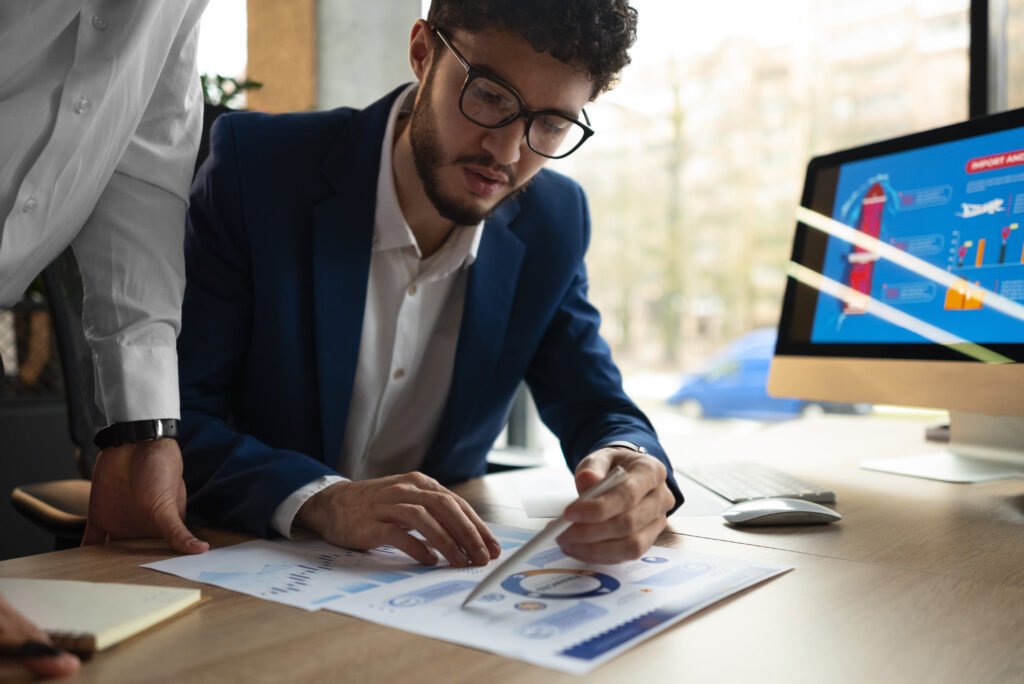 A young businessman wearing glasses and a blue suit is analyzing financial charts and graphs on a wooden desk. He is pointing at the document with a pen while another person, partially visible in a white shirt, is assisting. A computer screen in the background displays an infographic related to business or finance.