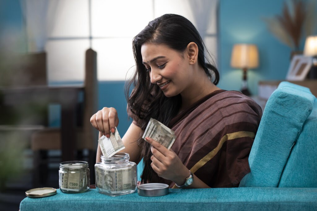 Smiling Indian woman in a saree sitting on a couch, placing ₹500 currency notes into a glass jar, symbolizing personal savings at home.