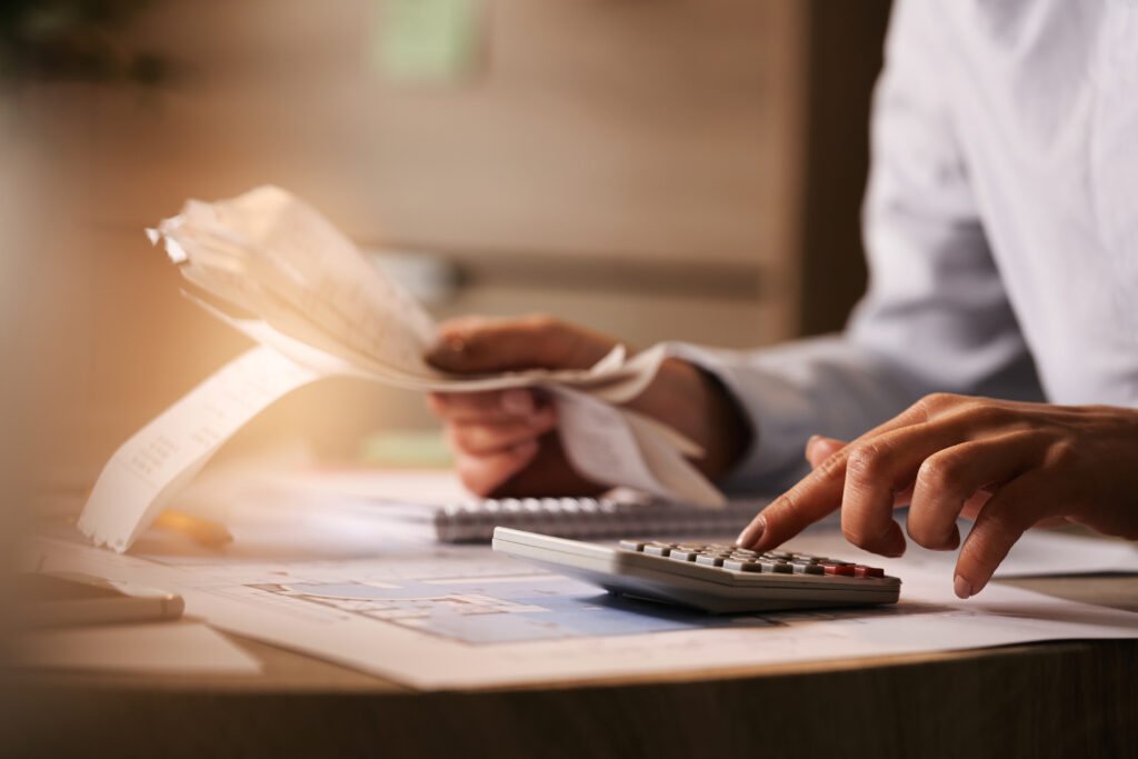 Close-up of a person's hands using a calculator and holding receipts while managing bills and taxes at a desk.