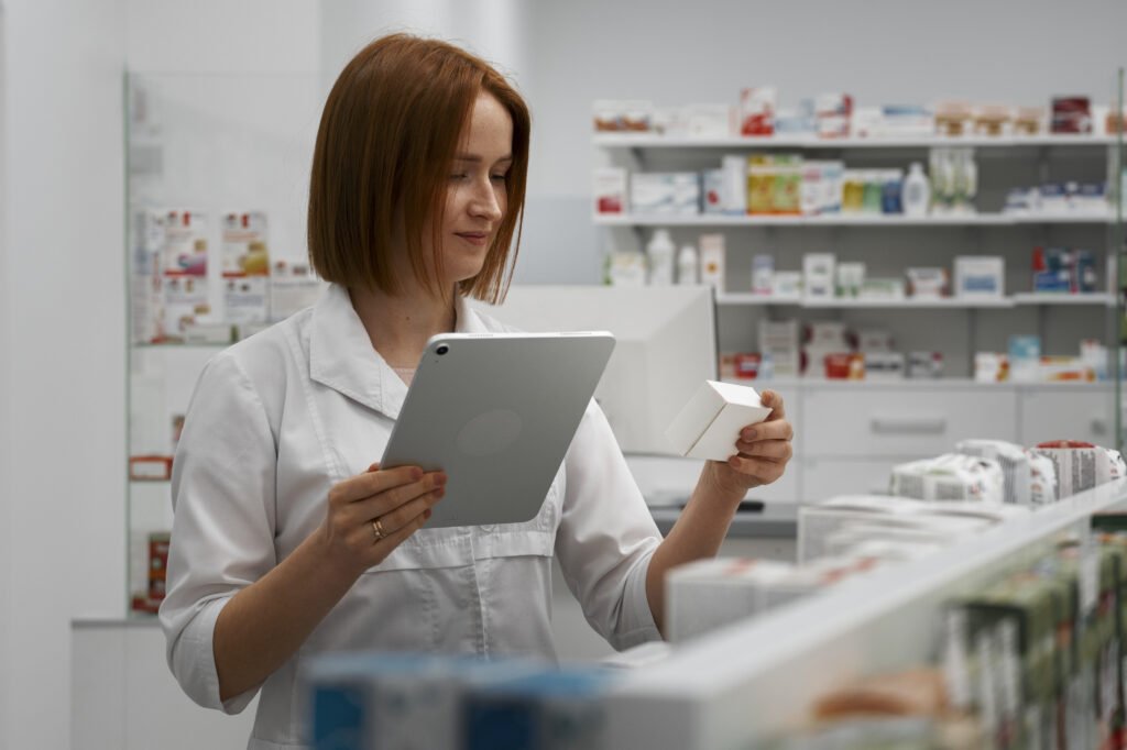 Female pharmacist in a white coat examining a medicine box while using a digital tablet in a modern pharmacy.