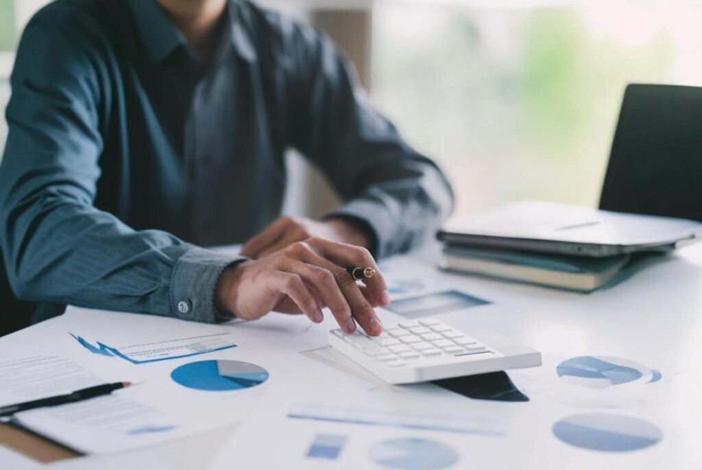 Businessman using a white calculator on a desk filled with financial reports, graphs, and documents, indicating financial planning.