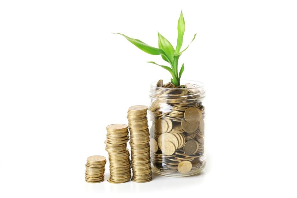 Stacks of gold coins beside a glass jar full of coins with a green plant growing out of it, symbolizing financial growth and savings.