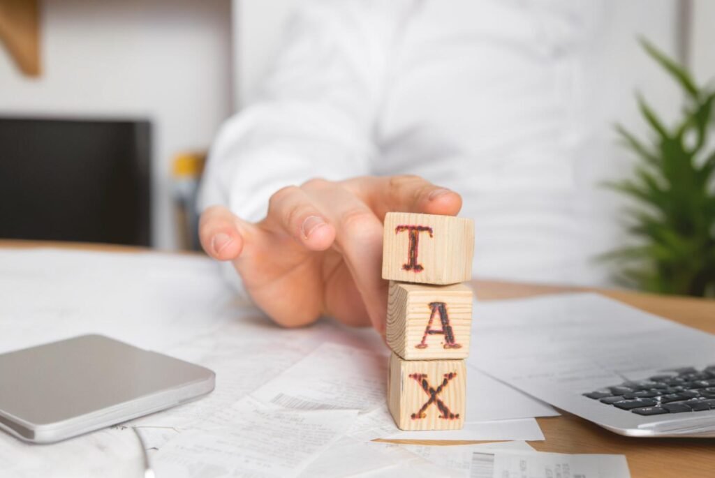 Close-up of wooden blocks spelling 'TAX' on a cluttered desk with financial papers, calculator, and a person in the background.