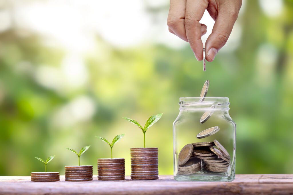 Hand dropping coins into a glass jar beside growing stacks of coins with small green plants, representing financial growth and savings investment.