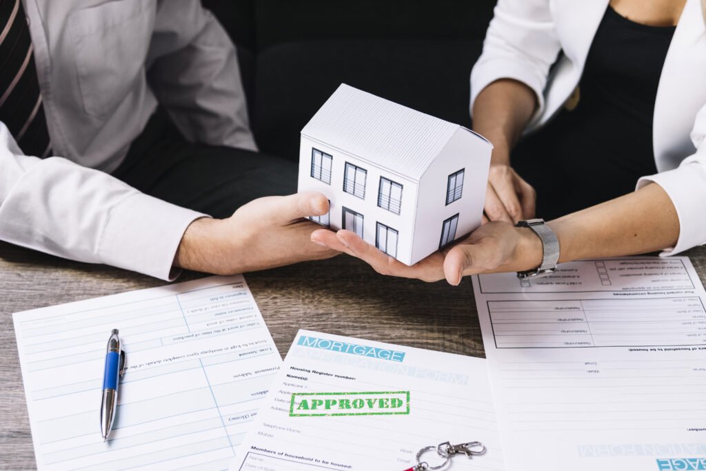 A real estate transaction, where a person in a white blazer hands over a small model house to another individual. The desk contains mortgage application forms, a pen, a pair of keys, and a document with a green "APPROVED" stamp.