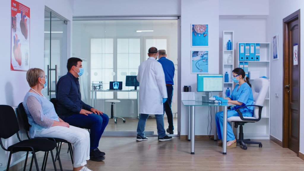 Patients wearing masks waiting in a hospital lobby while a nurse works on a computer and a doctor walks with a patient in the background.